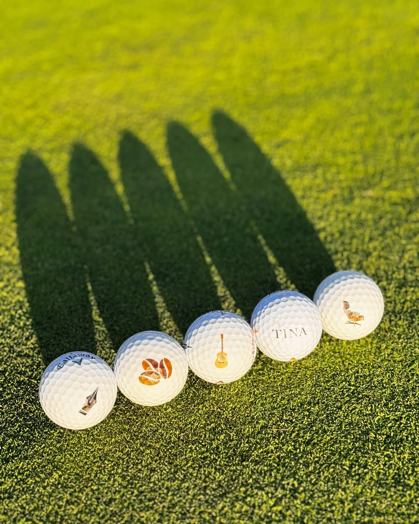 Five custom UV-printed golf balls in morning light on a putting green — sailboat, sunset, guitar, name and quail — by Print the Chicken in Orange County